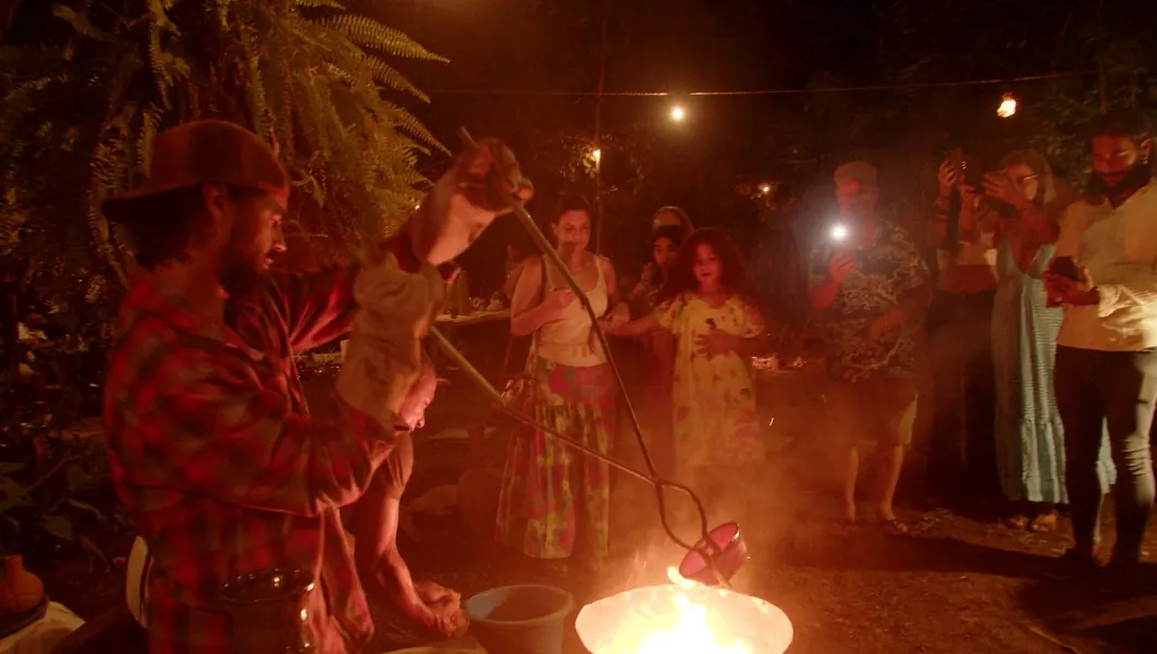 Pessoas reunidas à noite em volta de uma pequena fogueira, ao ar livre, sob luz alaranjada.