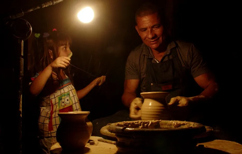Uma mulher de avental e um homem sorrindo modelam peças de barro sobre uma mesa, em um ateliê iluminado por uma lâmpada.