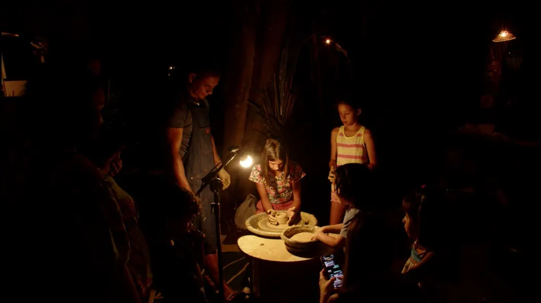 Pessoas ao redor de uma mesa, em ambiente escuro iluminado por uma luminária, durante uma oficina de cerâmica.