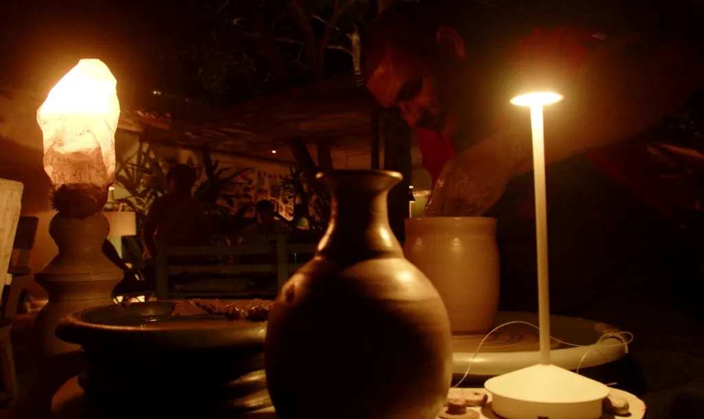 Mesa com um vaso escuro de cerâmica, uma pedra iluminada e um abajur de luz suave ao fundo.