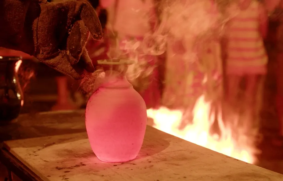 Vaso rosa incandescente ao lado de chamas, em uma queima de cerâmica ao ar livre; ao fundo, pessoas desfocadas.